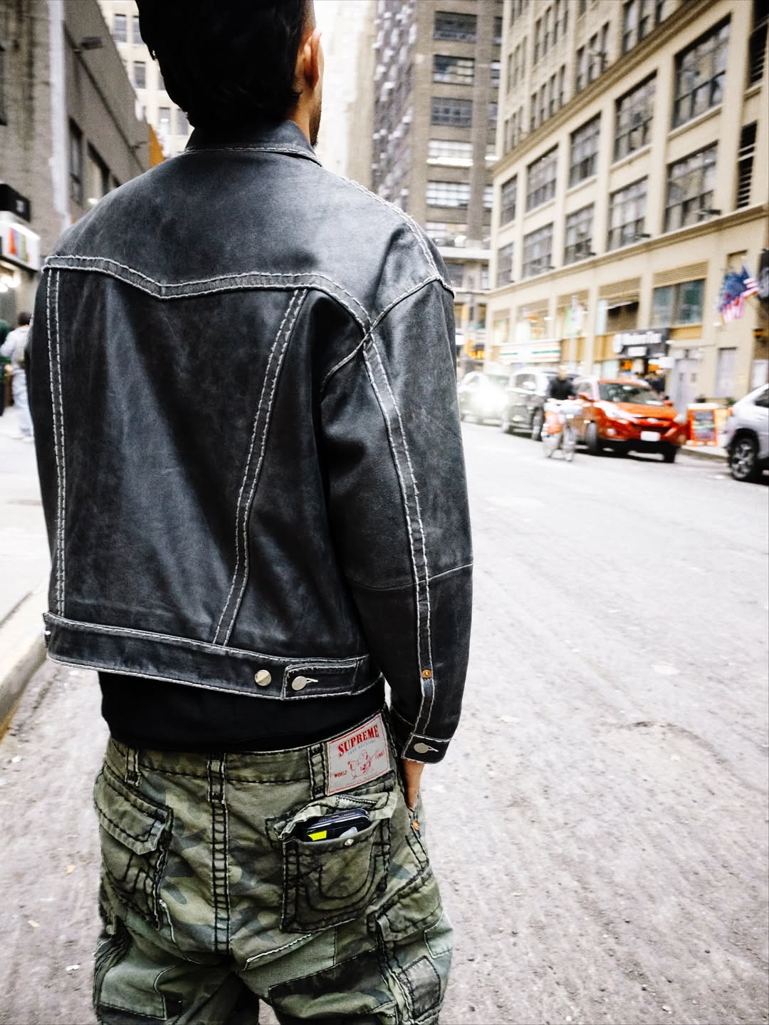 A model walks down a city street wearing a faded black Supreme x True Religion denim jacket paired with green cargo pants.