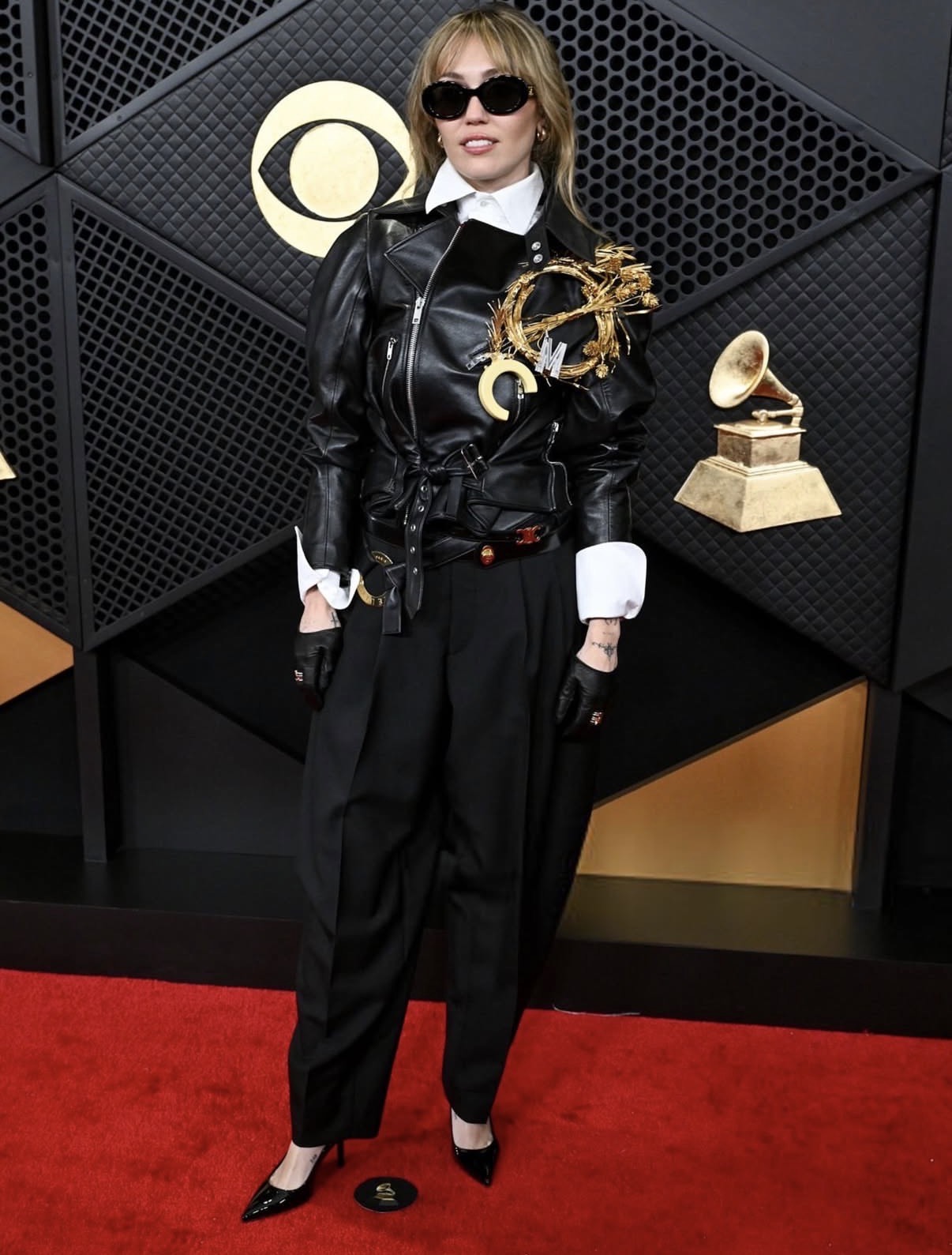 Miley Cyrus in a black leather moto jacket layered over a white button-down shirt with exposed French cuffs and structured collar details.