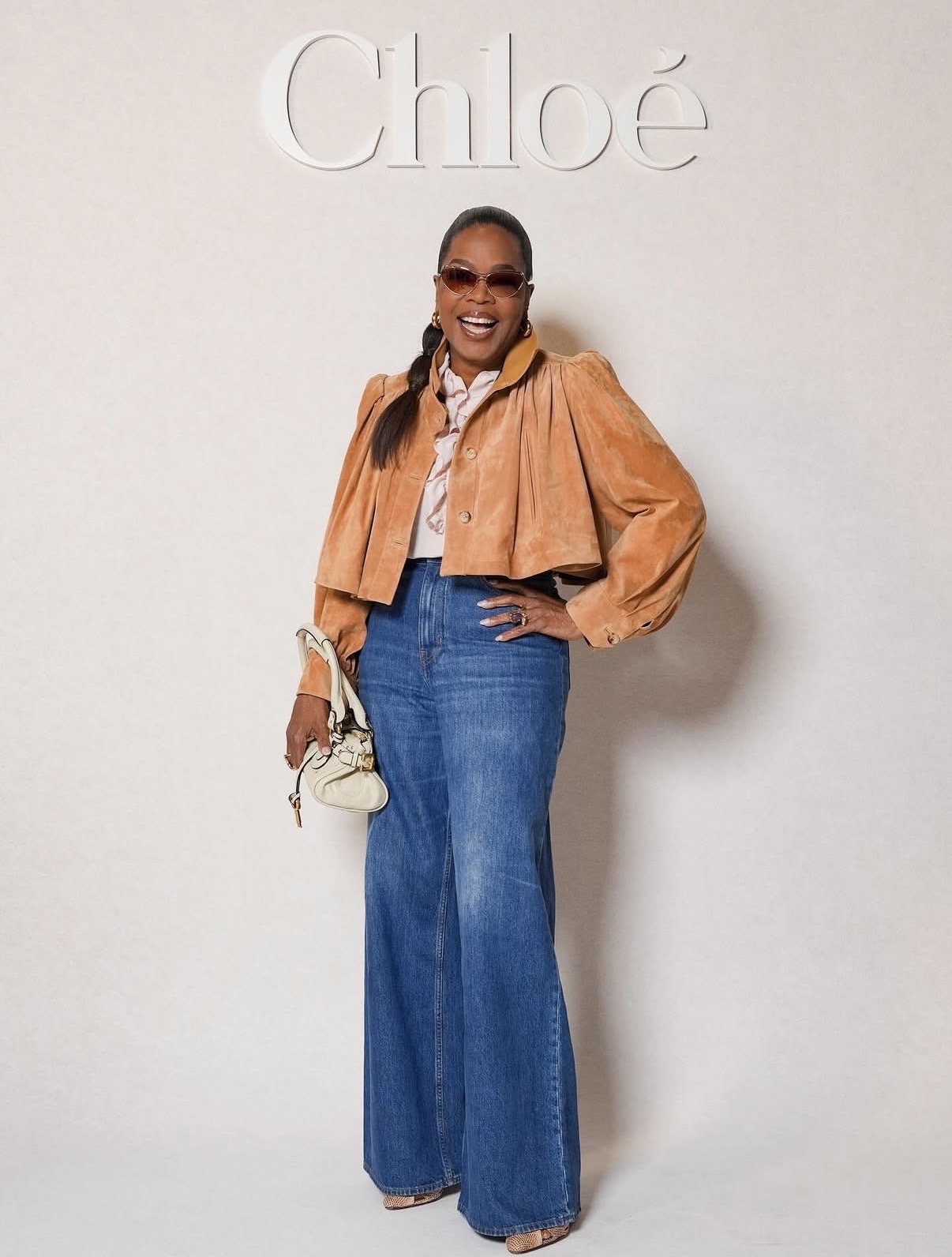 Oprah Winfrey in a cropped suede jacket, ruffled blouse, and wide-leg denim at the Chloé show during Paris Fashion Week.